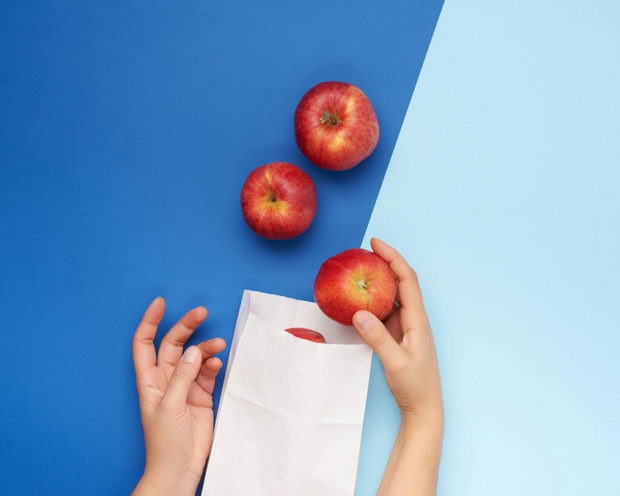 two female hands fold ripe red apples in a white paper bag, blue background